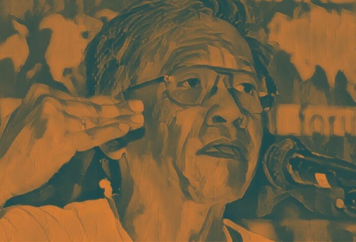 A close-up of Walden Bellow's face speaking and gesturing with a microphone. The photo has a paintlike orange filter overlayed.