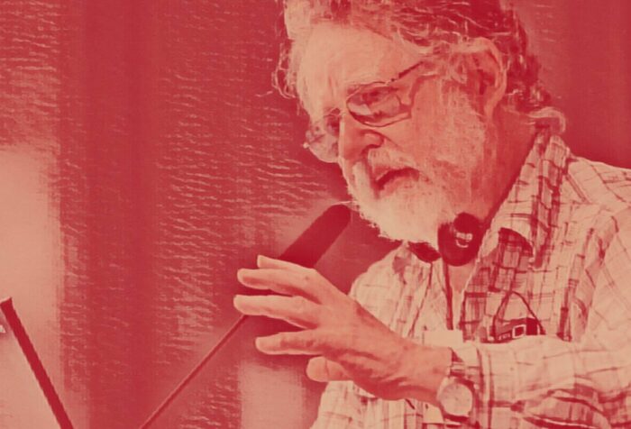 Edgardo Lander, a man with white facial hair and glasses, speaks at a microphone while gesturing with his hands. The photo has a red filter over it.