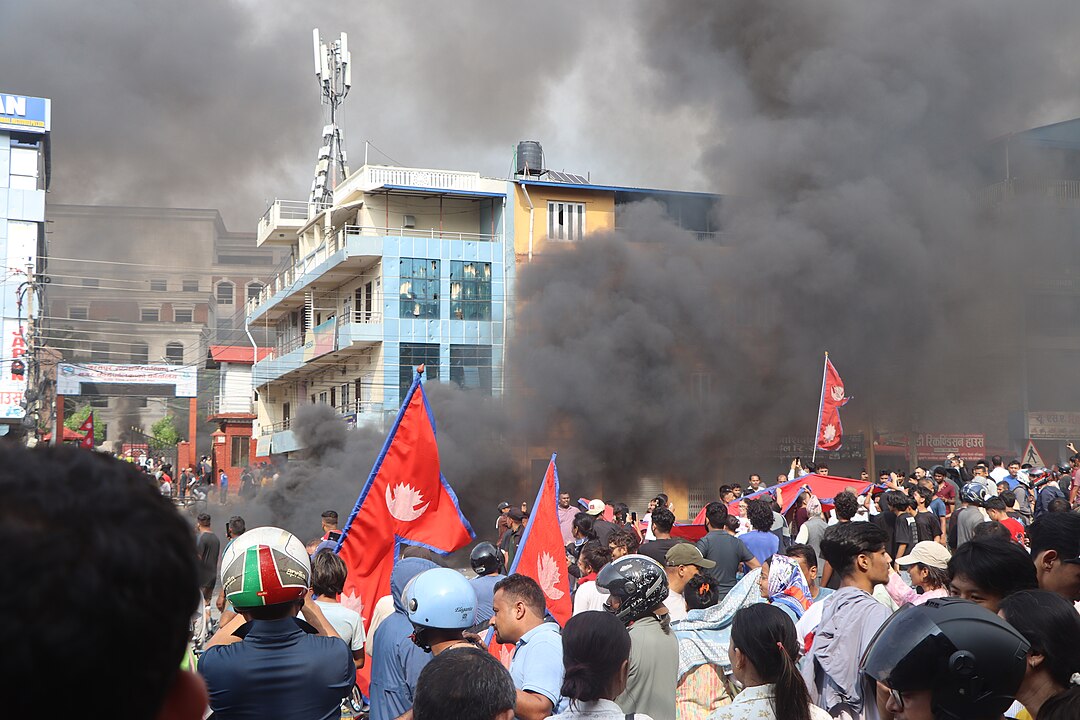 Photo of September 9, 2025 protests against corruption in bharatpur mahanagarpalika, Nepal. Photo shows large group of protestors, mainly younger, holding red flags, in a urban area in the midst of grey smoke from protest fires.