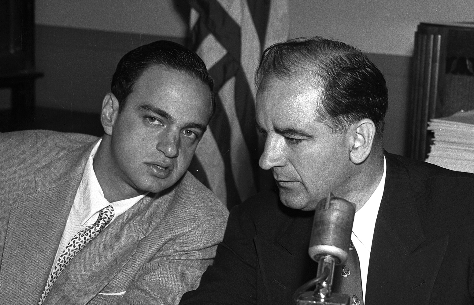 Black and white photo. Two men in suits lean together, talking.