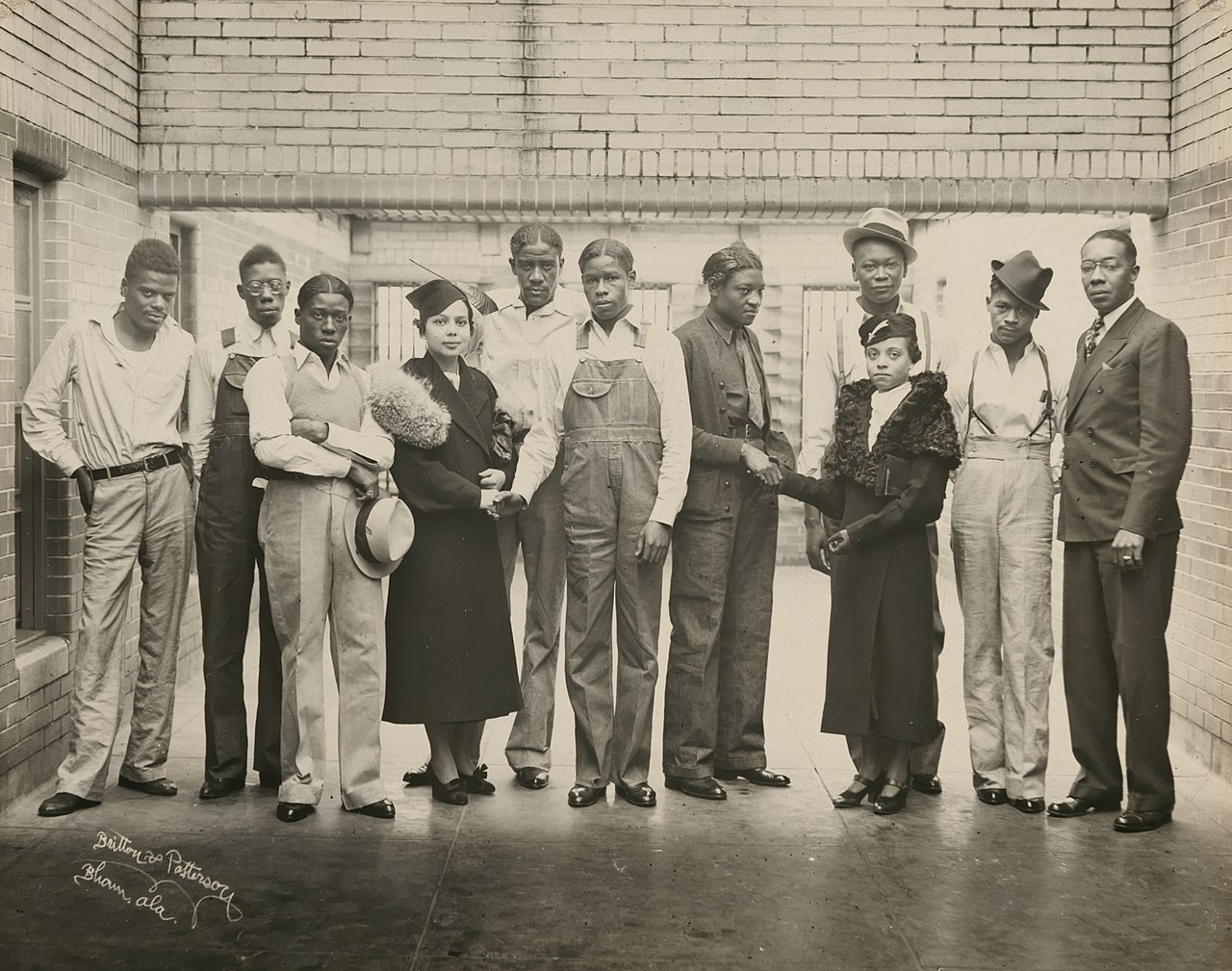 Black and white photo. Nine young Black men stand posing between brick walls with two young Black women.