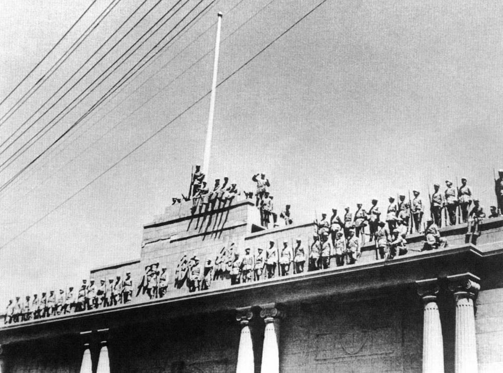 Black and white photo. Soldiers stand on the roof and ledges of a stone and concrete building.