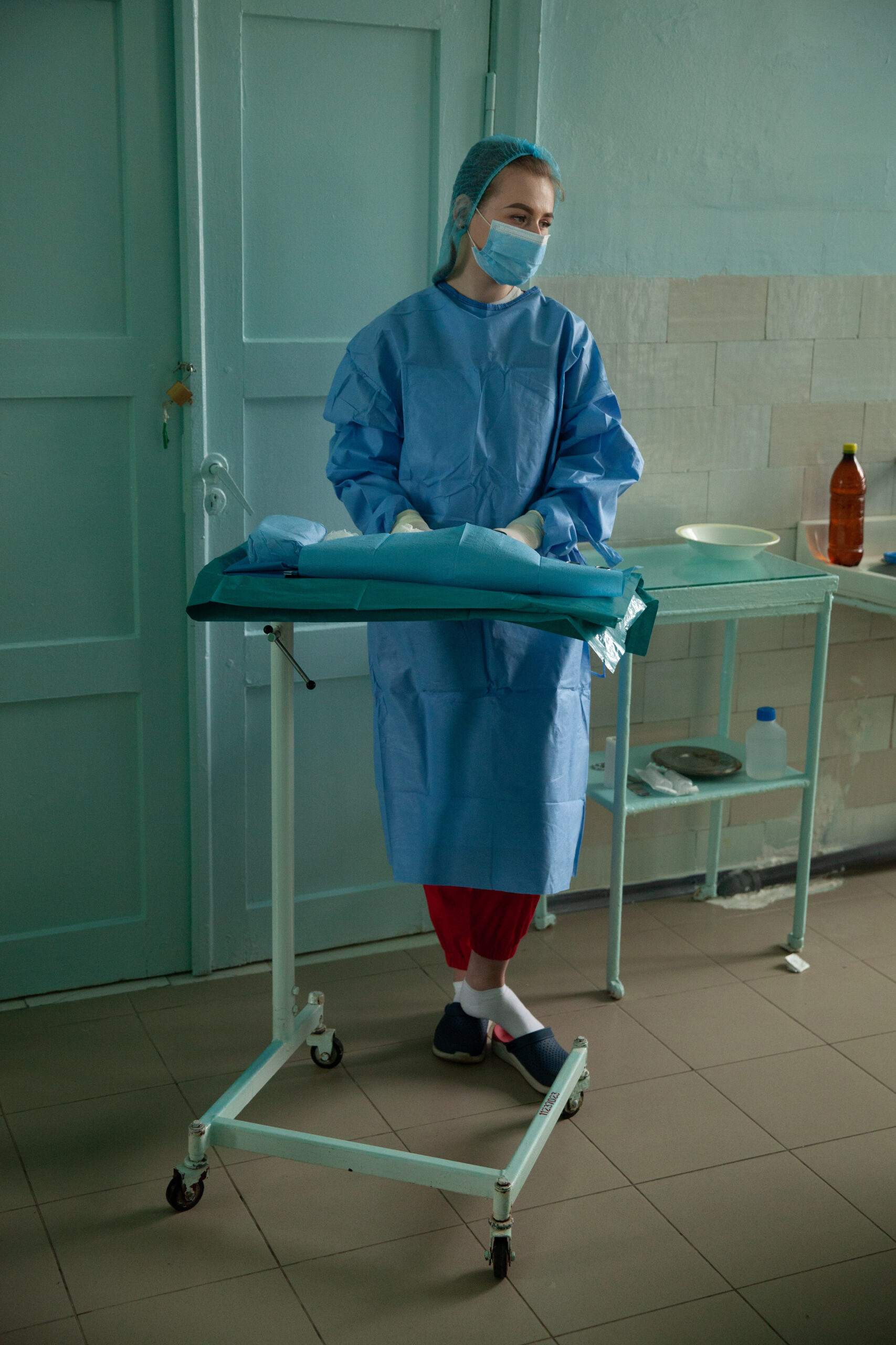 A nurse stands in blue scrubs, a mask, and a hair net, against a mint green wall.