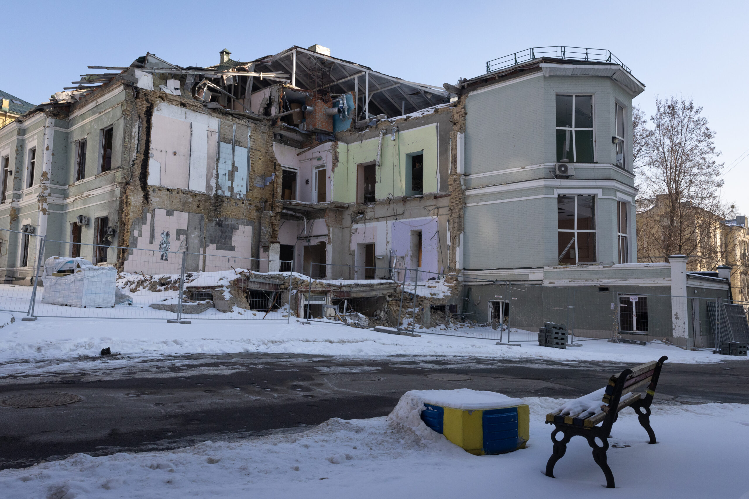 A photo of a set of dilapidated buildings behind metal fencing. In the foreground is a bench. There is snow on the ground.