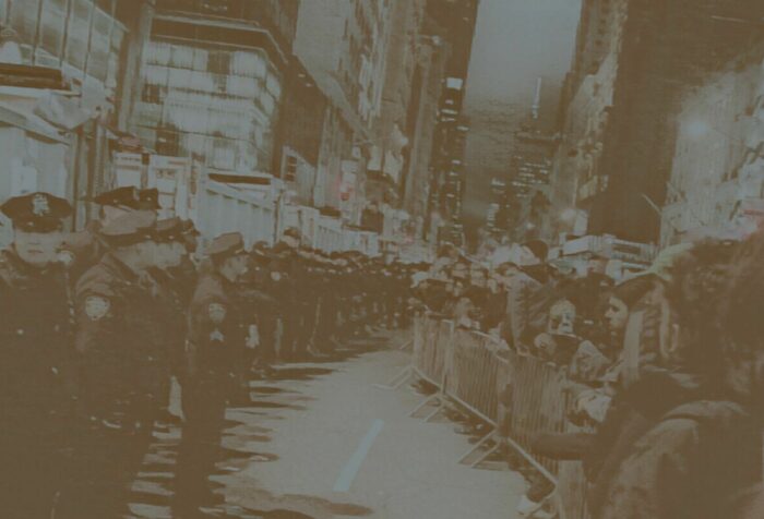 A photo of a line of police officers on the left, facing metal crowd barriers and a crowd of people behind them on the right, amidst city buildings. Photo is filtered in dark brown-grey.