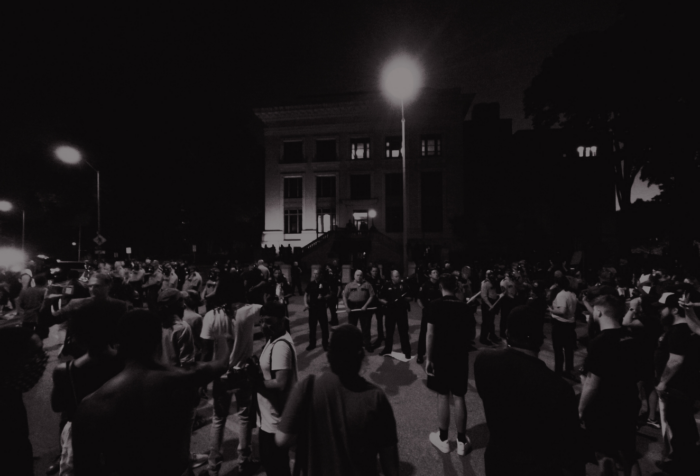 A black and white photo of a crowd facing off with a line of police officers in front of a building.