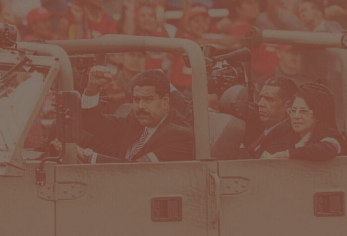 A photo of Nicolás Maduro driving an army car with others in it, holding his fist up, with a crowd of people behind. Photo is filtered with a faded brownish texture.