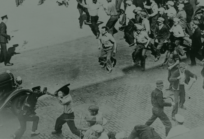 A black and white photo of a battle in the street between striking workers with pipes and police officers.