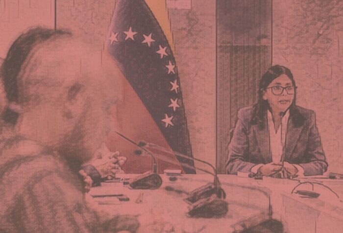 A photo of Delcy Rodriguez sitting at a table with other people, speaking with the Venezuelan flag behind her.