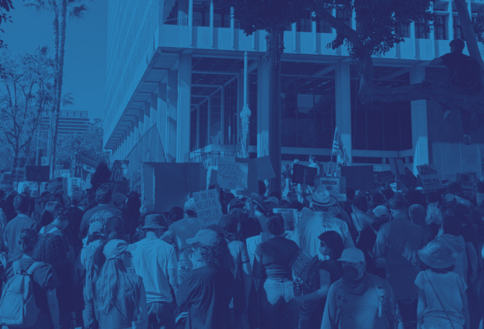 A blue-filtered photo of the back of a crowd holding anti-ICE signs in front of a building.