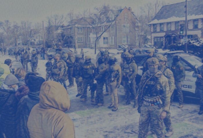 A blue-filtered photo of a line of ICE agents and a line of people facing each other at an intersection.
