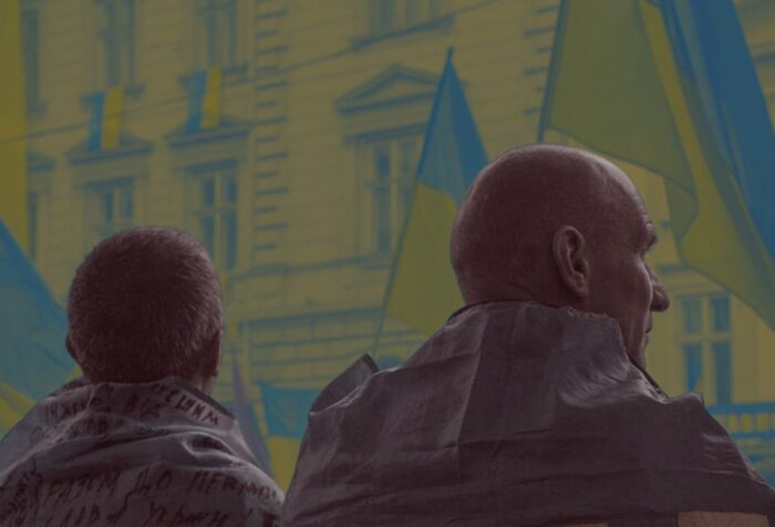 A photo of the backs of two people looking toward a building with Ukranian flags flying in front.