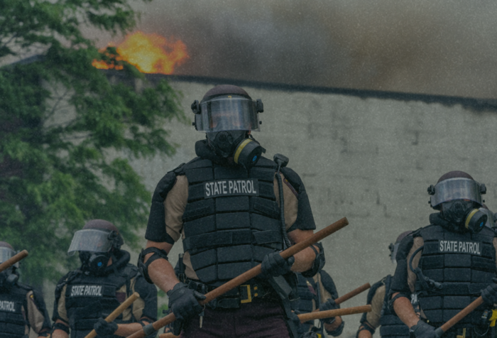A group of police officers with batons in front of a burning building.