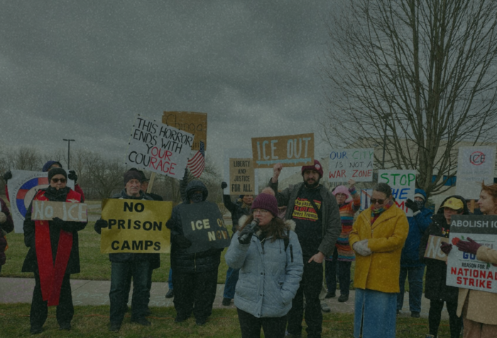 A crowd of people holding anti-ICE signs with one person speaking at a microphone.