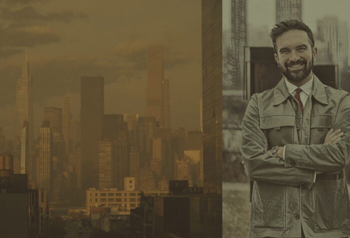 Zohran Mamdani stands with his arms crossed smiling in front of the NYC skyline.