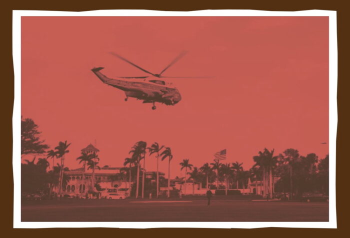 A red-tinted photo of a helicopter flying over palm trees and a US flag.