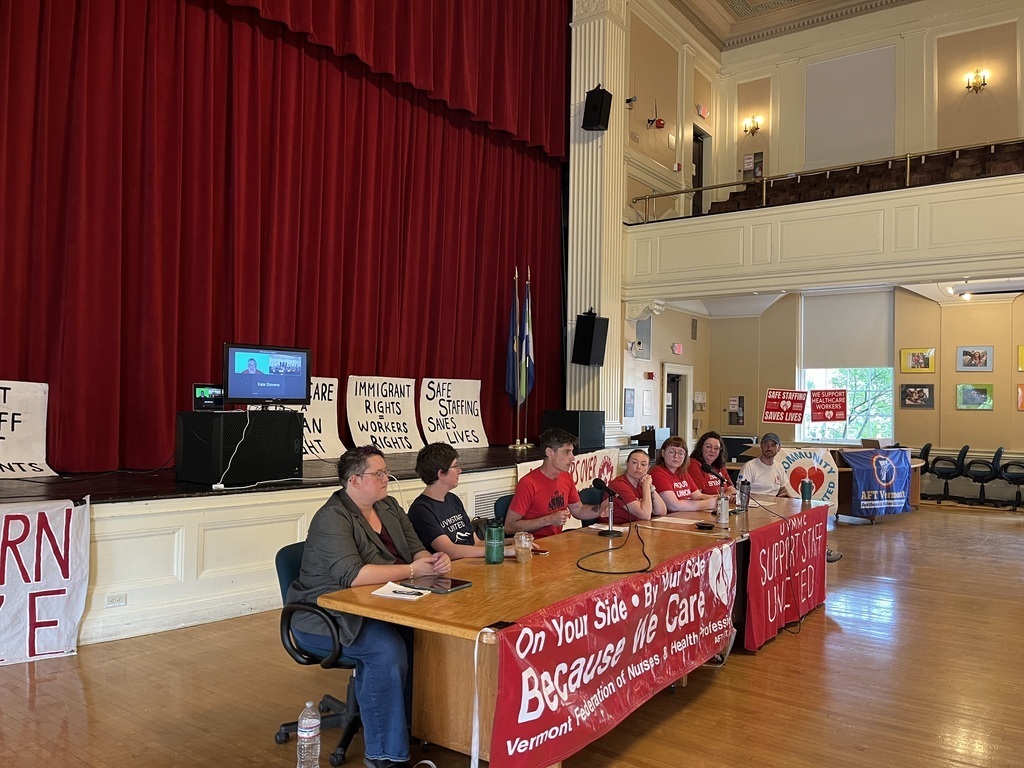 7 people sit at long table with red banner and signs about health care workers struggle.