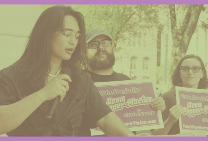 A photo of Rosa Pepper-Macias speaking into a microphone with people holding signs in the background.